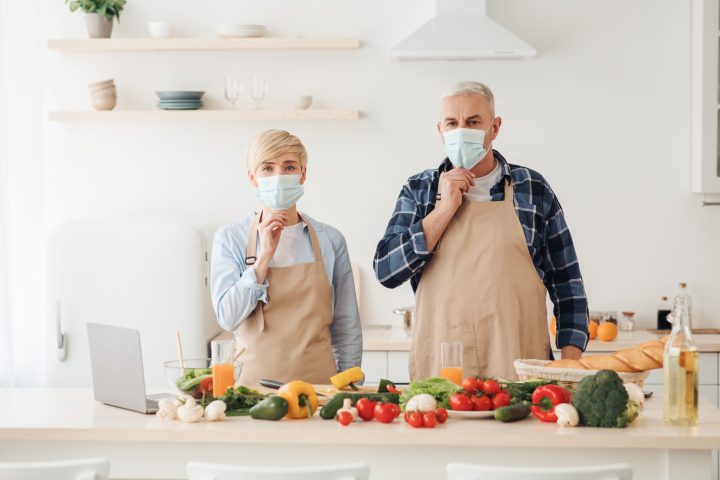 New normal, health care, be safety, modern food bloggers. Senior european man and wife in protective masks in modern kitchen interior with bright fresh vegetables on table and laptop, prepare meal